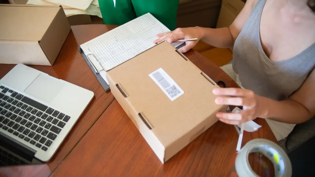 An ecommerce seller packing an order at a desk beside a laptop, the point where support volume starts outgrowing the founder