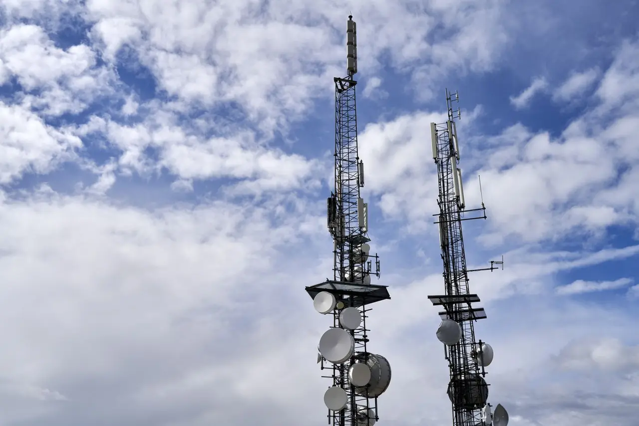 Telecommunication towers with antennas and dishes under a bright cloudy sky.