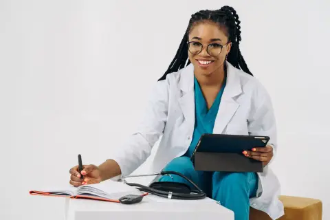 Smiling female doctor in scrubs with tablet and stethoscope, writing notes.