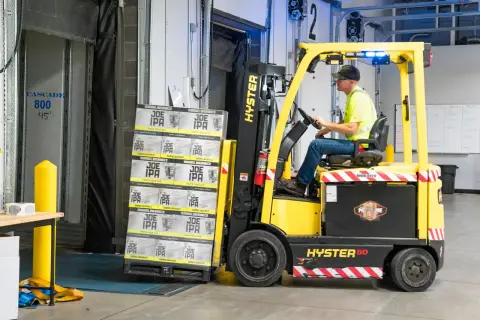 A worker drives a Hyster forklift moving Joe IPA boxes in a warehouse.